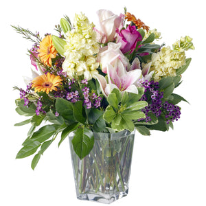 Garden arrangement featuring yellow gerbera daisies, white and pink lilies, pink roses, white stocks and purple wax flower arranged in Flower Studio signature garden style in a clear glass vase.
